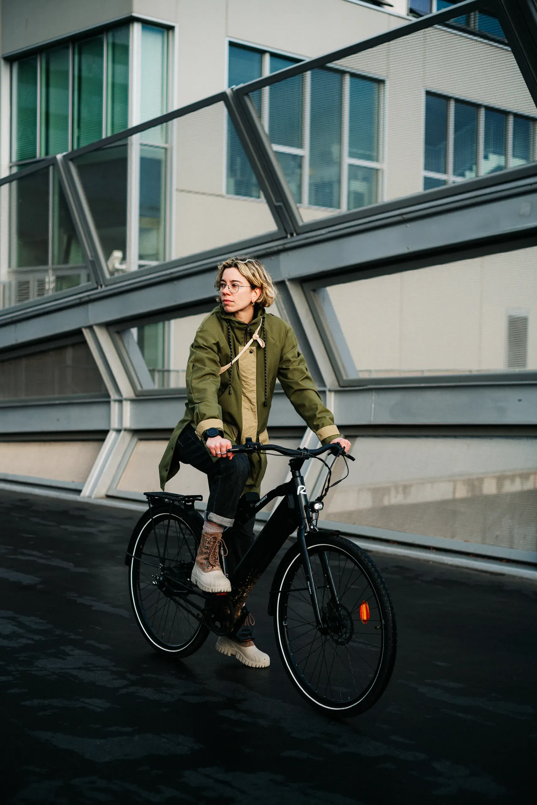 Une cycliste en parka kaki sur un vélo REF Bikes Urban Boost noir, sous une passerelle métallique